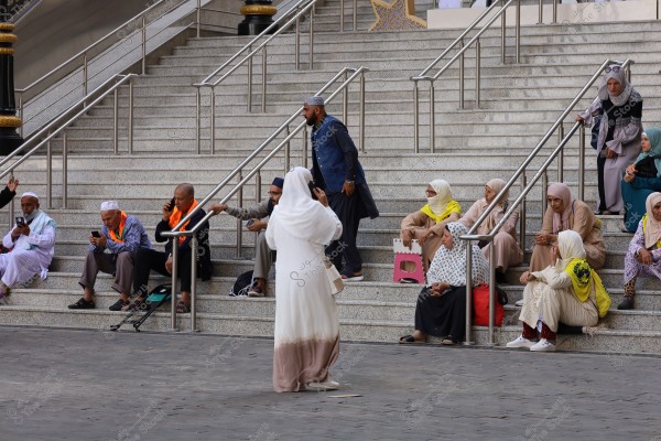 ** \r\nA group of people sitting on large, open stairs, with several individuals talking on mobile phones. A person in a white robe stands in the foreground. Many of the people are wearing traditional clothing, with some having headscarves. The scene has a calm, social atmosphere.\r\n\r\n**