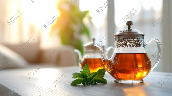 A glass teapot with silver handles and traditional Arabic motifs, filled with warm brown tea. The teapot has a silver lid, and next to it are green mint leaves on a light-colored wooden table. In the background, there is another similar teapot, and sunlight streams through a window, creating a warm and bright ambiance.