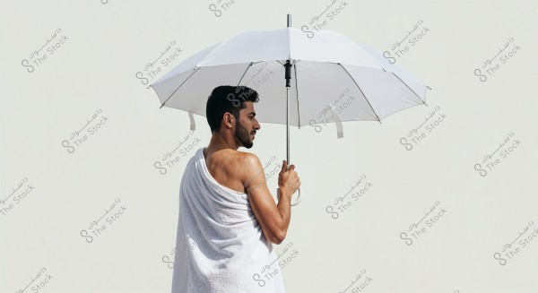 An image of a man standing against a white background, holding a white umbrella. The man is wearing a white robe and is shown from the back in a three-quarters angle. The image depicts him in a relaxed posture with a modern and minimalist appearance.