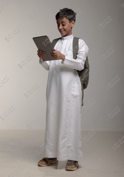 صورة لولد صغير يمسك جهازًا لوحيًا بيده, مرتديًا الزي التقليدي الأبيض المعروف بالثوب, وحقيبة ظهر سوداء. يبدو الولد مبتسمًا وينظر إلى الجهاز. الخلفية بسيطة وفاتحة اللون, مما يظهر تفاصيل الثوب والجهاز اللوحي بوضوح.
