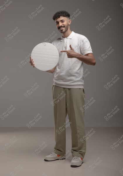 The image shows a man standing with a smile, wearing a white shirt and beige trousers. He holds a white circular disc and points at it with his other hand. The background is a soft gray.