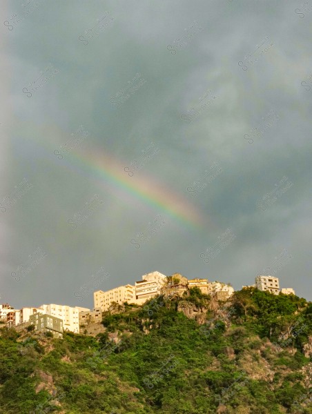 مشهد طبيعي لبلدة تقع على قمة جبل مغطى بالنباتات الخضراء, مع ظهور قوس قزح في السماء الملبدة بالغيوم. المباني ذات الطراز المعماري المتنوع تتراصف على حافة الجبل, مما يعطي إطلالة بانورامية.