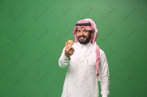 شاب سعودي على خلفية خضراء يرتدي الثوب و الشماغ، يقوم بعمل حركات بيده، يأكل الكعكة ، احتفالات العيد