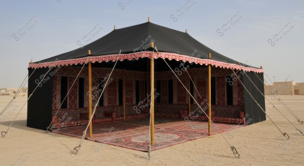 Image of a traditional Arabian tent made of black fabric, adorned with red and white traditional patterns along the edges. The tent is supported by ropes and wooden poles, situated in a sandy desert. Inside, there are decorative carpets, with some mud buildings visible in the distant background under a clear sky.