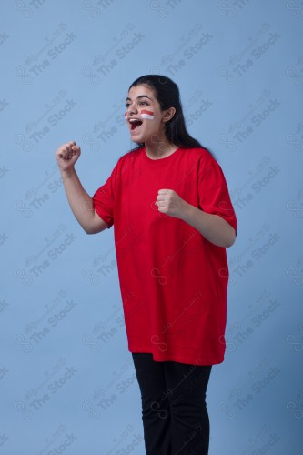 Portrait of a Saudi woman wearing a red T-shirt cheering the football team on a blue background and showing expressions of joy and enthusiasm, World Cup.