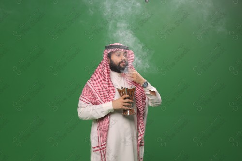 رجل سعودي يقف على خلفية خضراء يرتدي الثوب و الشماغ، يقوم بعمل حركات بيديه ، يبخر نفسه بالطيب و العود، احتفالات العيد