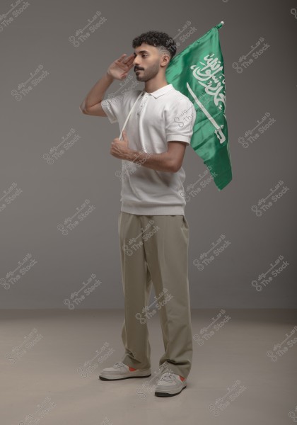 A man wearing a white polo shirt and beige trousers holds the flag of Saudi Arabia. He is positioned in a military salute. The background of the image is a simple gray.