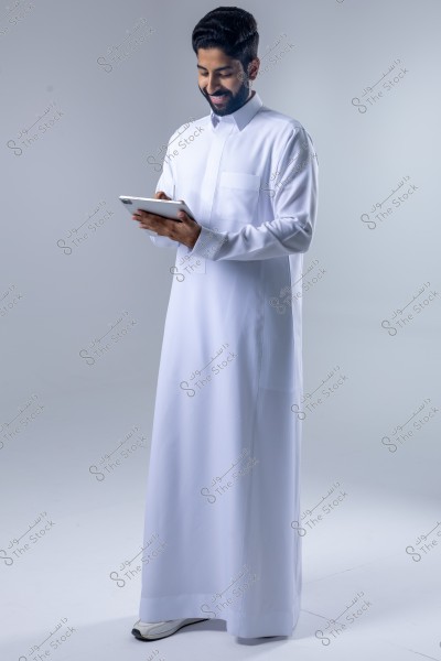 A portrait of a man standing in a softly lit studio, wearing a traditional white thobe, suggesting a Gulf origin such as Saudi Arabia. He is holding a tablet and smiling, conveying focus and ease.