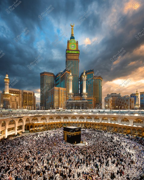 View of the Kaaba in the center of the Grand Mosque in Mecca, Saudi Arabia. Pilgrims dressed in white ihram garments gather around the Kaaba. In the background, the famous Mecca Clock Tower stands under a cloudy sky illuminated by the setting sun.