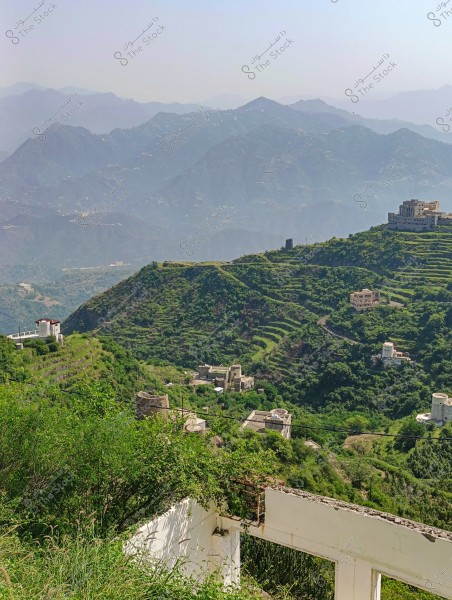 An image of a natural landscape showing a series of green, terraced mountains covered with dense vegetation. There are houses and buildings scattered across the hills, some highlighted by the bright sunlight. The scene extends to a range of mountains in the background, shrouded in a light mist that adds a sense of depth and tranquility.