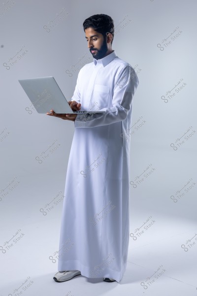 يظهر في الصورة رجل يقف في استوديو, يرتدي ثوبًا أبيض تقليديًا ويستخدم حاسوبًا محمولًا. يبدو في خلفية بيضاء ناعمة. الصورة تُظهر مظهرًا عصريًا وأنيقًا مع الإكسسوارات التكنولوجية.