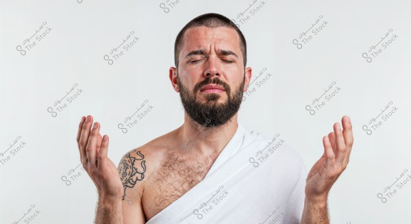 A portrait of a bearded man standing against a white background, wearing a white cloth draped over the right shoulder, with a prominent tattoo on the left shoulder. His facial expression is neutral as he closes his eyes and slightly raises his hands as if in meditation or prayer.