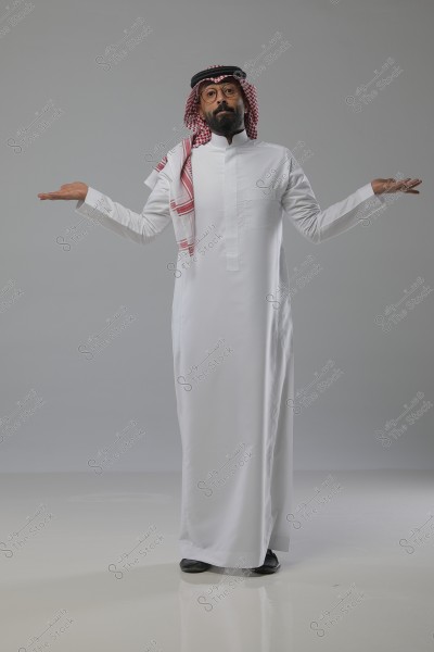 Image of a man standing in a studio, wearing a traditional Saudi thobe in white with a red ghutra and agal. The man is raising his hands in a gesture that suggests questioning or puzzlement.
