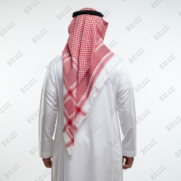 Image of a man from behind wearing a white thobe, a red and white ghutra, and a black agal. The clothing indicates traditional Saudi attire. The background is plain white.