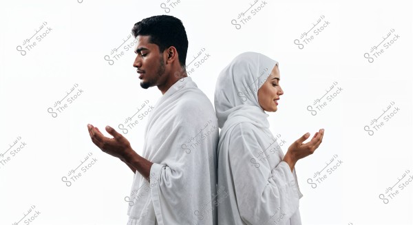 The image shows a man and a woman standing back-to-back. Both are dressed in white Ihram clothing, with the man wearing traditional Ihram garments while the woman wears a headscarf covering her hair and a white abaya. They both appear focused and reverent, with their hands raised in a prayer position. The background is entirely white, highlighting the details of their attire and stance.