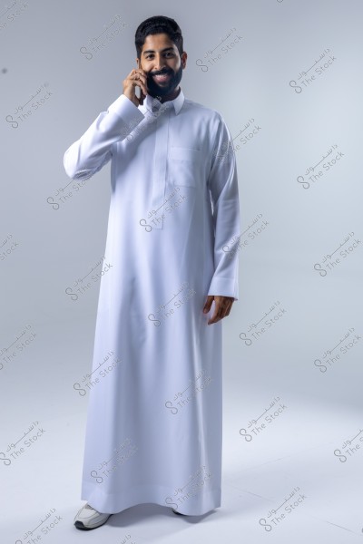 An image of a man standing in traditional Gulf attire, wearing a white thobe and smiling while talking on the phone. He is set against a neutral background, with good lighting highlighting the details of his clothing.