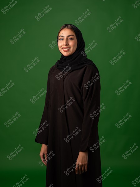 امرأة ترتدي حجابا وعباءة سوداء تقف أمام خلفية خضراء.