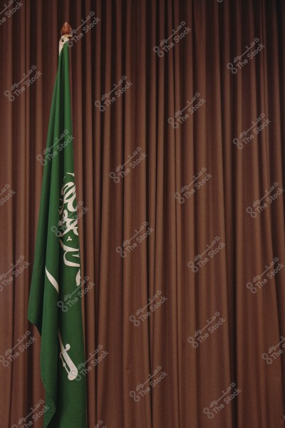 Image of the Saudi Arabian flag standing next to a brown curtain. The green flag features the Shahada and a sword in white.