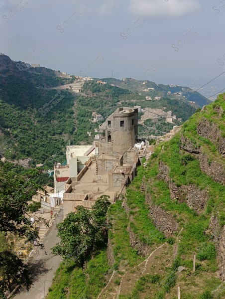 A scenic view of a lush mountainous area. The image shows residential buildings on a hilltop, surrounded by green terrain and valleys. The sky is clear with a few clouds. A narrow road leads to the buildings and runs parallel to the mountain.