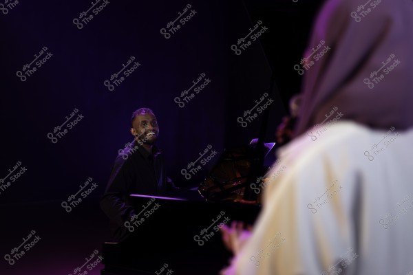 عازف بيانو يبتسم على المسرح بينما يقف شخص آخر في المقدمة, الأضواء خافتة في الخلفية.