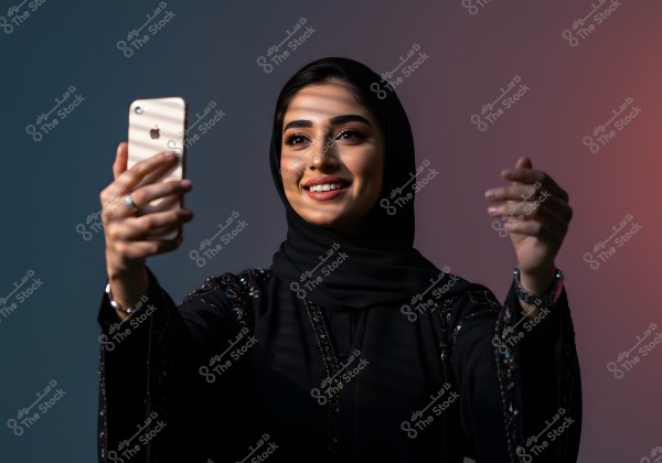 Image of a woman wearing a black abaya taking a selfie with a gold phone. The woman appears happy and smiling, with light reflections on her face. The background is a blend of dark blue and pink.