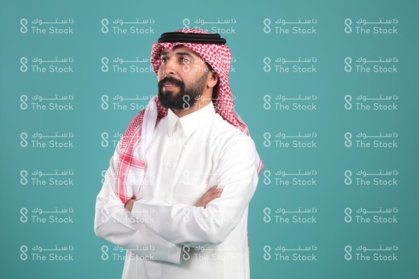 بورتريه رجل سعودية يرتدي الثوب ويقف أمام خلفية تركوازية ، تعابير وجه تدل على التساؤل والحيرة والتفكير ، لحظة اتخاذ قرار ، صورة شخصية