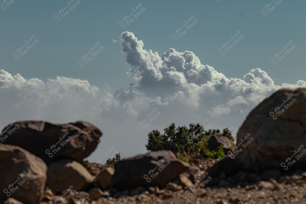A landscape image showing large, scattered clouds in a clear sky, with a prominent cloud in the center. In the foreground, there are large rocks on the ground with small green patches indicating plants and bushes in the background.