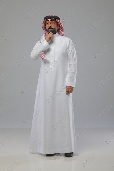 A man standing pensively, wearing a white thobe with a red checkered shemagh and a black agal. He is wearing glasses and has his hand on his chin as if in thought. The background is gray.