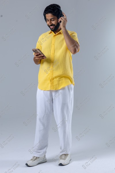 A young man stands in a studio with a smiling expression, wearing a yellow shirt and white trousers, with headphones on his ears, holding a smartphone. He appears relaxed and seems to be enjoying music.