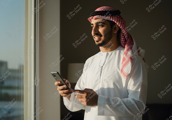 A portrait of a man standing by a window holding a smartphone. He is wearing traditional Gulf attire, including a white thobe and a red and white checkered headscarf called a keffiyeh. He appears to be smiling and looking outside as sunlight illuminates his face.