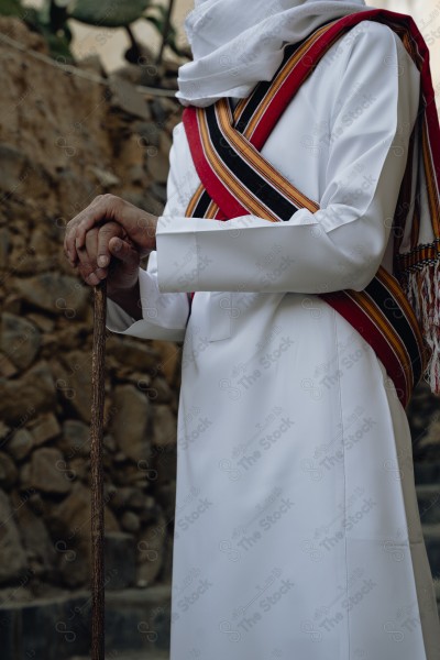 A snapshot of a Saudi man wearing traditional costume on the founding day, standing in front of a mud building and leaning on a stick, traditional costume, foundation day, ancient ancient buildings