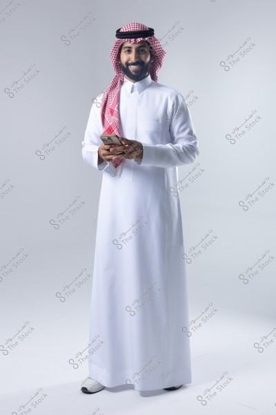صورة لرجل بزي سعودي تقليدي, يقف مبتسمًا ويحمل هاتفًا في يده. يرتدي ثوبًا أبيض وغترة حمراء وبيضاء مع عقال أسود. الخلفية بيضاء وبسيطة.