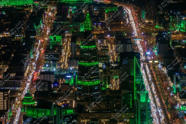 Nighttime image of a city illuminated with green lights, showing an aerial view of Riyadh, Saudi Arabia. The picture features several tall buildings and busy roads filled with cars whose headlights create streams of light. The prominent building with a distinctive design, known for its green lighting, stands out in the center of the image.