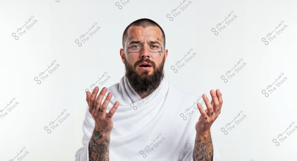 A portrait of a man with a short beard and tattoos on his face and hands, wearing a simple white robe draped over his shoulders, showing an expression of confusion or inquiry. The background of the image is pure white.
