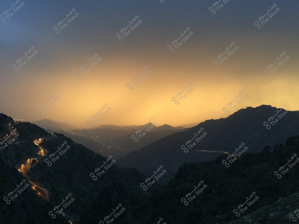 Image shows a sunset view over a mountain range, with a glowing horizon in shades of orange and yellow. There is a winding road illuminated by street lights, harmonizing with the surrounding mountains and valley views.