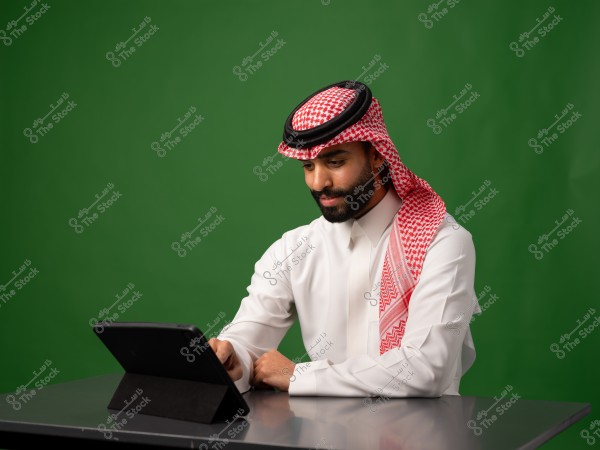 رجل يرتدي الزي الخليجي التقليدي ويستخدم جهاز لوحي على طاولة, مع خلفية خضراء.