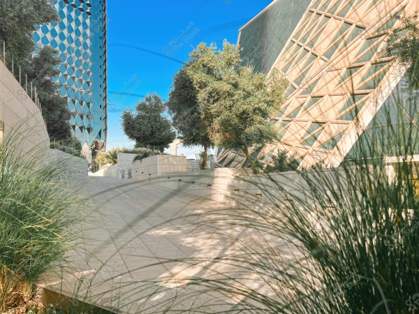 An image of a pathway next to two modern architectural buildings, with the first featuring a reflective glass facade in a geometric pattern, and the second showing a slanted exterior with triangular decorations. The buildings are surrounded by green trees and foliage along the pathway, with a clear blue sky in the background.