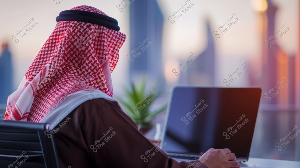 An image of a man wearing a brown thobe and a headband on a red and white shemagh, sitting in front of a laptop in an office with a city view. A glass window behind him shows modern buildings and a sunset scene, with a green plant on the desk.