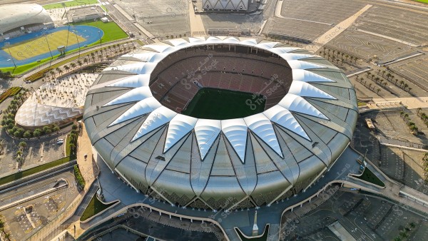 Aerial view of a large sports stadium featuring a circular design and a distinctive metallic roof resembling a crown. The green field is visible in the center, surrounded by spectator seating. Expansive parking lots and other sporting facilities surround it, with pathways and neatly lined palm trees.