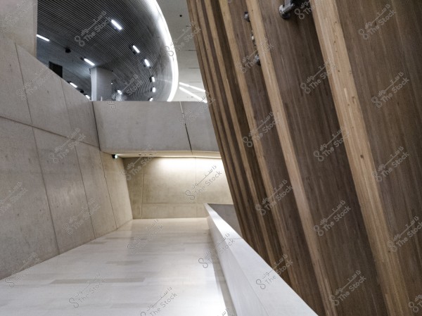An image showing an interior space with a modern architectural design. The walls are angled and made of concrete with wooden panels on the right side. The curved ceiling features integrated lighting, providing a smooth flow of light. The floor is light-colored polished stone, creating a sense of simplicity and calm.