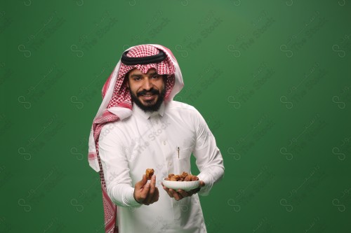 بورتريه رجل سعودي يرتدي الزي السعودي التقليدي يحمل طبق من التمر ويقدم تمرة لأحدهم