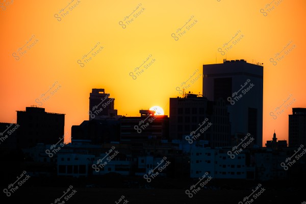 View of a sunset behind city buildings, showing the city skyline silhouetted against a warm orange sky.