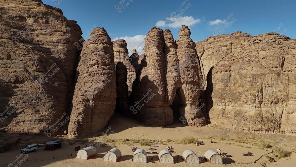 A natural landscape of a desert valley with large and varied rock formations under a blue sky. In the foreground, there is a group of white tents and vehicles on the sandy area below the rock formations. Tables and chairs are visible next to the tents, giving a sense of a tourist camping site.