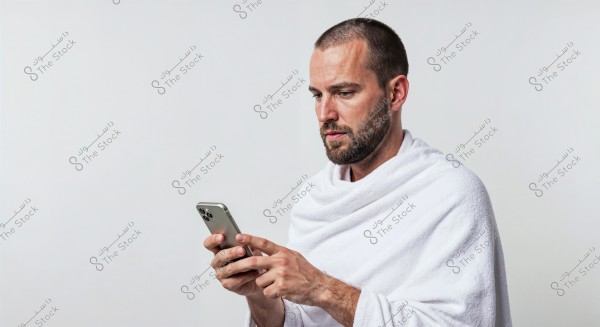 A portrait of a man focused on a smartphone in his hands, wearing a simple white garment resembling Ihram clothing. The background is white, and his expression is one of concentration and engagement with the screen.