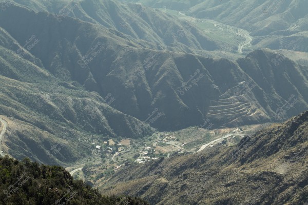 A scenic view of a mountainous valley area with green hills, showing rolling mountainous terrain, small villages, and farms. The road winds around the mountains, and terraced fields are visible on the mountain slopes.
