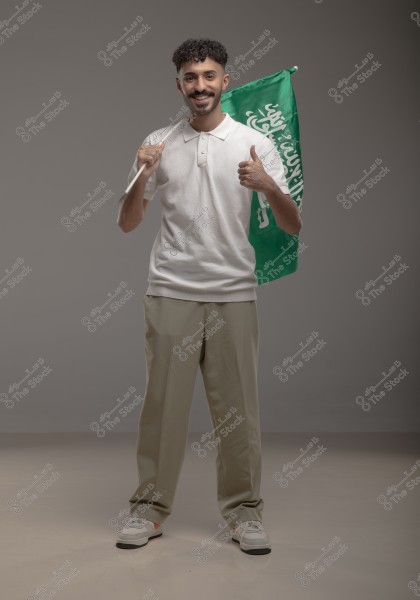 A man standing and smiling while holding a green flag, wearing a white shirt, beige pants, and sneakers. A gray wall is in the background.