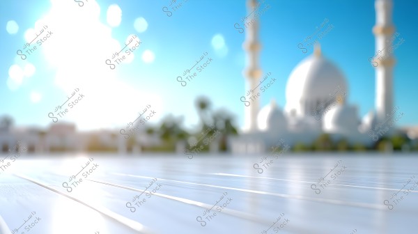 A blurred view of a large mosque with white domes and tall minarets under a clear blue sky in the background. The foreground shows a shiny surface reflecting bright sunlight.