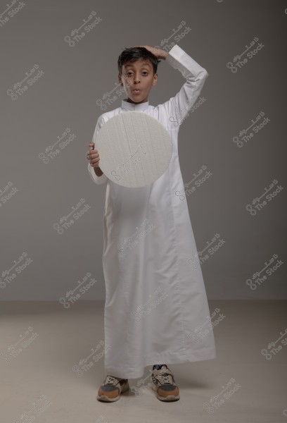 A photo of a child wearing a traditional white thobe, holding a white circle in his right hand, with an expression of surprise or astonishment on his face. The child stands against a gray background and is wearing sneakers.