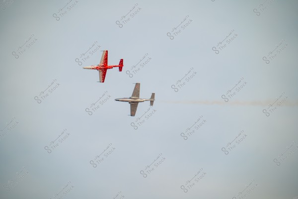 Two aircraft flying in a clear blue sky. The top aircraft is red with a white and red tree pattern, while the lower aircraft is grey with some markings on the tail. A smoke trail follows the lower aircraft.
