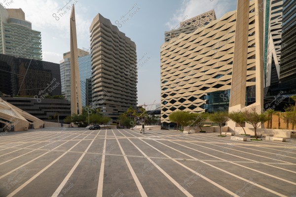 A large plaza surrounded by modern, futuristic architectural buildings. Tall structures featuring unique geometric designs, some with sharp angles and glass facades, stand prominently. Tall pillars add an aesthetic touch to the space. Small trees are sporadically placed along the plaza, which is tiled with a linear pattern.